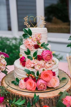 a white wedding cake with pink flowers and greenery on top is sitting on a tree stump