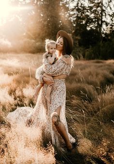 a woman holding a baby in her arms while walking through tall grass with trees in the background