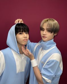 two young men standing next to each other in front of a red wall wearing blue and white hoodies