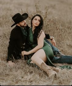 a man and woman sitting on the ground in a field with tall grass, wearing cowboy hats