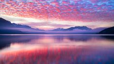 colorful clouds are reflected in the still water at sunset on a lake with mountains in the background