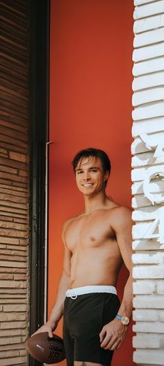 a shirtless man holding a football in front of a red wall with white shutters