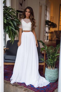 a woman wearing a white two piece dress standing in front of a potted plant