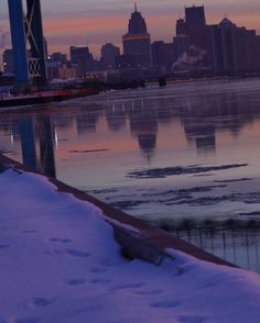 the city skyline is reflected in the icy water at dusk, with snow on the ground