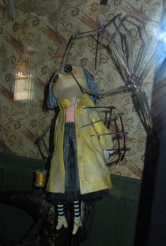 a stuffed animal wearing a yellow coat and black dress standing next to a wallpapered wall