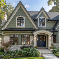 a gray house with two story windows and black shutters on the front door is surrounded by greenery