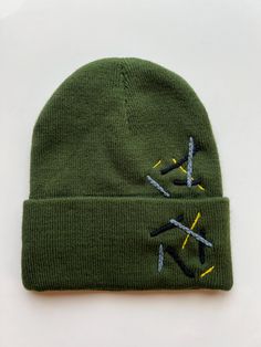 Army green embellished beanie with blue, black, and yellow accents. Trendy Green Fitted Hat, Green Beanie For Cold Weather, Trendy Green Hat For Fall, Green Casual Winter Beanie, Green Winter Beanie Cap, Fitted Warm Green Hat, Green Beanie Cap For Winter, Green Beanie Cap For Streetwear, Green Winter Beanie One Size