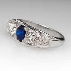 This lovely circa 1930s antique sapphire ring is centered with one (1), bead set, oval cut natural blue sapphire weighing 0.81 carats and flanked to each side by one (1), bead set, old European cut diamond. The shoulders of the ring are each bead set with two (2) round single cut diamonds. The gemstones are bordered with milgrain edging. Engraved details accent the side faces of the ring. The ring measures 7.3mm at the top, rises 3.7mm above the finger, tapering to 2.9mm wide and 1.6mm thick at the base of the shank. The ring is currently a size 6 and we offer complimentary resizing to fit. Classic Sapphire Birthstone Ring, Classic Oval Sapphire Ring, Classic Sapphire Halo Ring, Antique Oval Three Stone Diamond Ring, Vintage Three Stone Sapphire Ring, Classic Sapphire Ring With Lab-created Gemstone, Classic Blue Sapphire Ring, Classic Oval Sapphire Ring With Rose Cut Diamonds, Vintage Sapphire Three Stone Diamond Ring