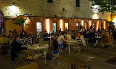 people are sitting at tables in an outdoor cafe with bright lights on the side of the building