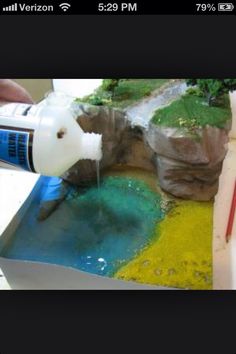 someone is pouring water into a small pond with rocks and trees on it, while another person holds a paintbrush in their hand