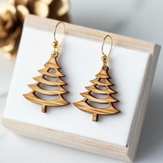 two wooden christmas trees are hanging from gold earwires on a white box next to a golden ornament