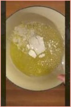 a white bowl filled with yellow liquid on top of a wooden table next to a person's hand