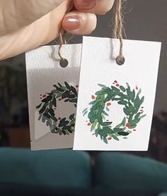 someone holding up two christmas cards with holly wreaths on them, one is white and the other is green