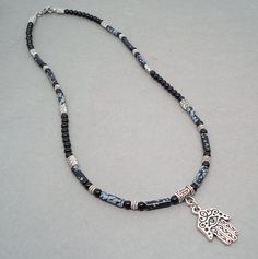 "This distinctive necklace features a metal pendant of Hamsa, known as an amulet shaped like a hand and is thought to protect against the evil eye. This detailed Hamsa charm is a made-in-USA high quality TierraCast product, which is made of pewter in antiqued fine silver finish. It hangs from a necklace that I designed with natural Snowflake Obsidian stone tube beads in black with whitish gray patches resembling snowflakes, and Black Onyx gemstone round beads. I have also added antiqued silver f Black Amulet Necklaces With Polished Beads, Black Amulet Necklace With Polished Beads, Black Amulet Style Necklaces With Polished Beads, Black Amulet Beaded Necklaces For Jewelry Making, Hand Of Hamsa, Hamsa Charm, Mens Necklace, Obsidian Stone, Snowflake Obsidian