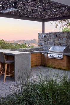 an outdoor kitchen with grill and bar stools under a pergolated roof at sunset