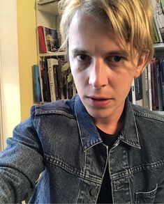 a young man with blonde hair wearing a denim jacket and looking at the camera while sitting in front of a bookshelf