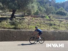 a man riding a bike down a street next to a stone wall and tree covered hillside