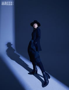 a woman in a black coat and hat standing on a white floor with her shadow