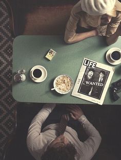 two people sitting at a green table with coffee and magazines on it, one person has his head down