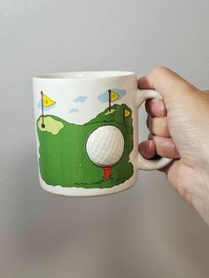 a hand holding a coffee mug with a golf ball on it and the hole in the cup