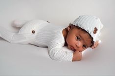 a baby laying on its back wearing a white knitted outfit with buttons and a hat