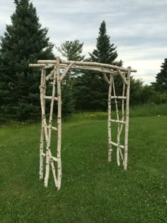 a wooden arch made out of branches in the middle of a grass field with trees behind it