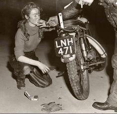 a woman kneeling down next to a man on a motorbike while he fixes the tire