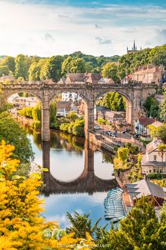 an old stone bridge over a river in the middle of town with trees around it