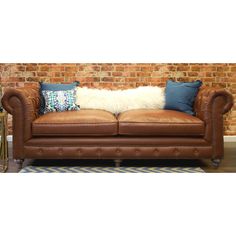 a brown leather couch with blue and white throw pillows on it's back, in front of a brick wall