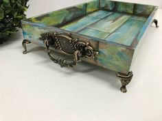 an ornately painted wooden tray with metal handles on a white table next to a potted plant