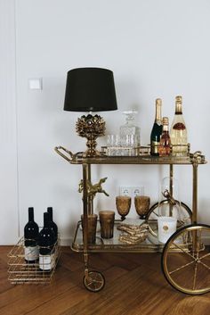 a gold bar cart with wine glasses and liquor bottles sitting on top of it next to a lamp