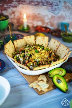 a bowl filled with food sitting on top of a table next to crackers and an avocado