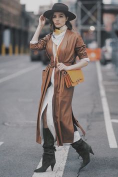 a woman is walking down the street wearing a brown coat and black hat with her hand on her hip