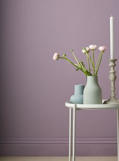 two vases with flowers on a table in front of a purple wall and a white candle