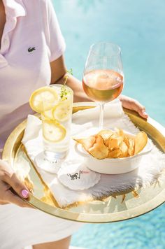 a woman holding a tray with food and wine on it next to a pool in the background