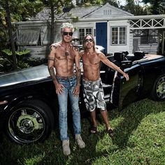 two shirtless men standing next to a black car in front of a white house