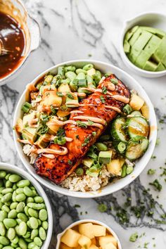 two bowls filled with salmon and vegetables next to green beans, avocado wedges and sauce