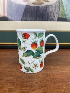 a white coffee cup with strawberries on it sitting on a table next to a painting