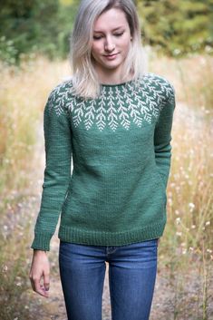 a woman wearing a green sweater and jeans standing in front of tall grass with her hands in her pockets