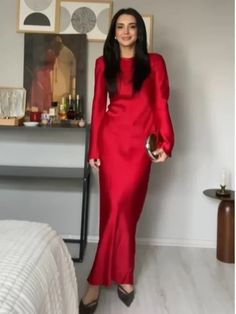 a woman in a long red dress standing next to a bed and looking at the camera