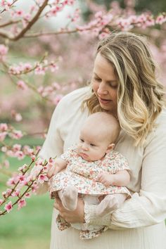 a woman holding a baby in her arms while standing next to a tree with pink flowers