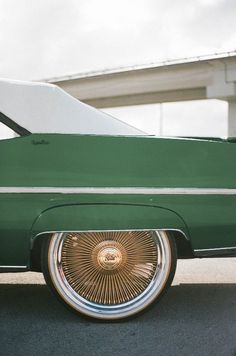 an old green car with chrome rims parked on the side of the road in front of a bridge