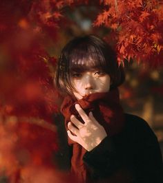 a woman is standing in front of a tree with red leaves on it and she has her hands wrapped around her neck