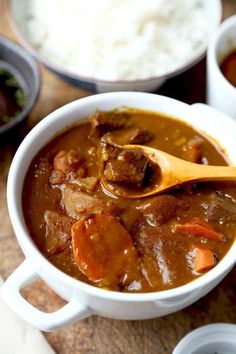 a white bowl filled with meat and carrots on top of a wooden table next to a spoon