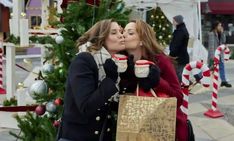 two women kissing each other in front of a christmas tree while holding coffee mugs