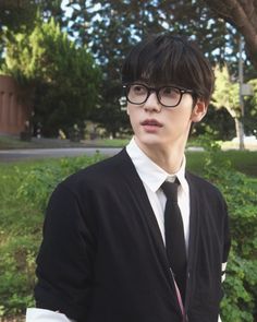 a young man wearing glasses and a black cardigan standing in front of some bushes