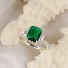 an emerald and diamond ring sitting on top of a white flowery cloth with two diamonds around it