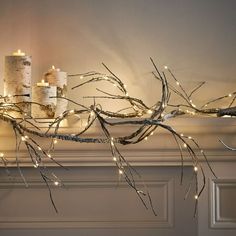 some candles are lit up on a mantle with branches and lights hanging from the top