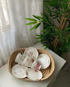 a wicker basket filled with teacups and saucers next to a potted plant