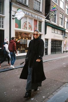 a woman is standing on the sidewalk in front of a store wearing sunglasses and a black coat